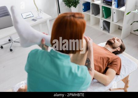 manipulateur manuel flou fléchissant la jambe blessée de l'homme tout en faisant le massage de soulagement de douleur dans le centre de réadaptation, image de stock Banque D'Images