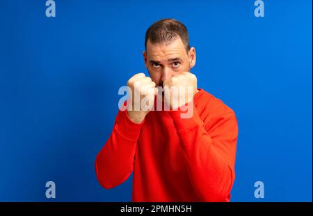 Homme hispanique barbu dans son 40s portant un maillot rouge debout garde prêt à commencer un combat avec ses poings, isolé sur fond bleu. Banque D'Images