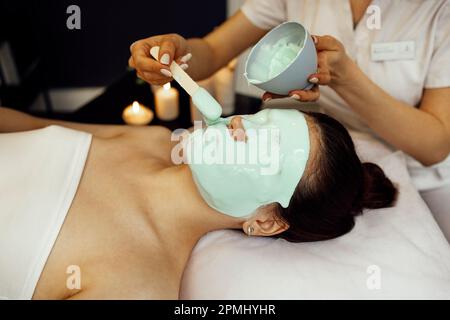 Soin de la peau du visage. Belle jeune femme recevant un masque cosmétique dans le salon de beauté. Esthéticienne appliquant le masque d'alginate sur le visage de la femme avec le Sod lisse Banque D'Images