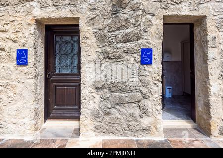Deux pictogrammes bleus marquent des toilettes intégrées dans un vieux mur en pierre naturelle d’un bâtiment de l’Université Arkin des Arts Créatifs et du Design (ARUCAD) à Kyrenia (turc : Girne), République Turque de Chypre du Nord (RTNC). Une lourde porte en bois ornée de fer forgé et une seconde porte ouverte encadrent le mur. Fondée en 2017 par le groupe Arkın, l’université est une institution thématique dédiée exclusivement à l’art, au design et à la communication. Banque D'Images