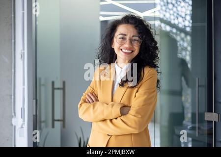 Portrait d'une femme d'affaires réussie à l'intérieur du bureau, patron latino-américain en costume jaune souriant et regardant l'appareil photo, femme mature avec cheveux bouclés debout avec les bras croisés. Banque D'Images