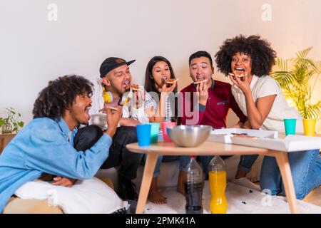 Un groupe d'amis multiethniques joyeux sur un canapé en train de manger une pizza à la maison Banque D'Images