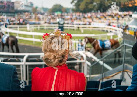 Femme en robe rouge portant un beau fascinateur regardant les chevaux défiler avant la course Banque D'Images