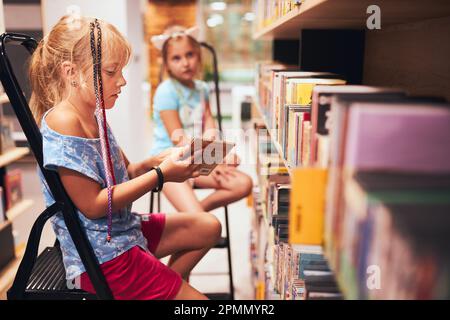 Écolières à la recherche de livres audio dans la bibliothèque de l'école. Étudiants choisissant des livres. L'éducation élémentaire. Faire des devoirs. Apprendre à partir de livres. Retour à sch Banque D'Images