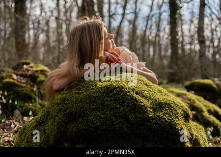 Snowdrops galanthus blond. Une fille en robe blanche s'est couché sur une pierre dans la mousse dans un pré avec des gouttes de neige dans une forêt de printemps Banque D'Images