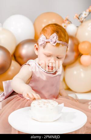 Petite fille à tête rouge fête son premier anniversaire. Cake crash smash, les mains manger. fête de balons familiaux de 1 ans. Séance photo professionnelle. Studio photo Banque D'Images