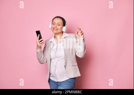 Bonne femme enceinte charmante dans un casque sans fil, tenant un téléphone mobile, dansant, isolée sur fond rose Banque D'Images