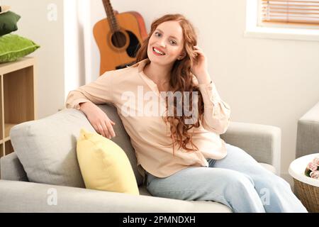 Belle femme à tête rouge assise sur un canapé à la maison Banque D'Images