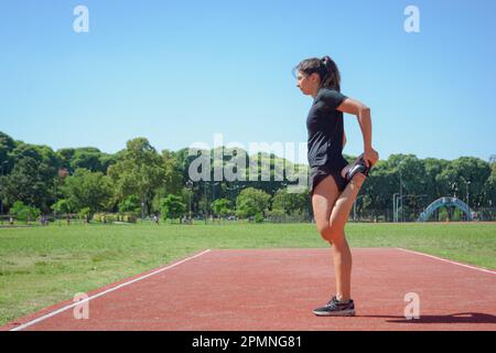 vue latérale de la jeune femme latine argentine, vêtue de vêtements noirs, debout sur la piste de course qui étire le muscle fémoral de la jambe gauche, avec des arbres et des sk Banque D'Images