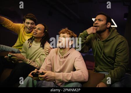 KIEV, UKRAINE - 13 FÉVRIER 2023: L'homme indien focalisé assis près d'amis interraciaux gais jouant le jeu vidéo dans le club de jeu, image de stock Banque D'Images