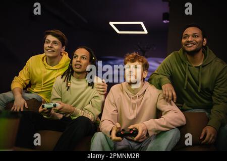 KIEV, UKRAINE - 13 FÉVRIER 2023: Souriants hommes multiethniques assis près d'amis avec des joysticks dans le club de jeu, image de stock Banque D'Images