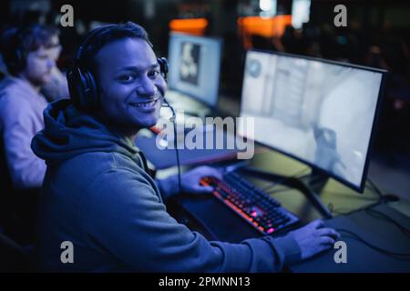 Souriant gamer indien dans le casque regardant l'appareil photo près de l'ordinateur dans le cyber club avec l'éclairage, image de stock Banque D'Images