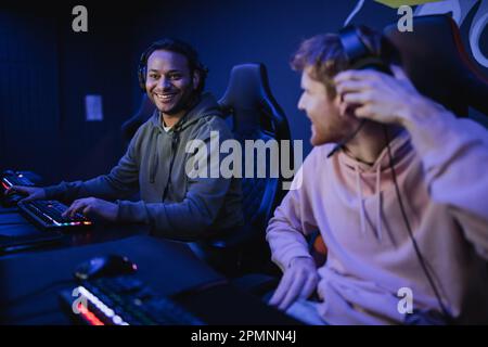 Gamer indien souriant dans un casque assis près d'un ami flou dans un cyber-club, image de stock Banque D'Images