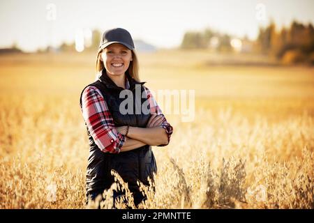 Portrait d'une fermière mature debout dans un champ de céréales, posant pour la caméra pendant la récolte au coucher du soleil ; Alcomdale, Alberta, Canada Banque D'Images