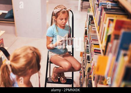 Écolières à la recherche de livres audio dans la bibliothèque de l'école. Étudiants choisissant des livres de jeu. L'éducation élémentaire. Faire des devoirs. Apprendre à partir de livres. Retour à Banque D'Images