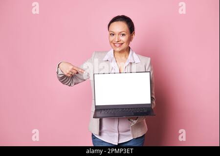 La femme enceinte qui travaille sourit à l'appareil photo, pointe son doigt sur l'écran numérique blanc de l'ordinateur portable maquette. Grossesse et carrière Banque D'Images