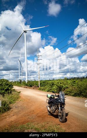 Une grosse moto noire garée sur une piste de terre à côté d'une ligne de transmission de puissance, d'énormes éoliennes et une plantation de café dans la province de Gia Lai Banque D'Images