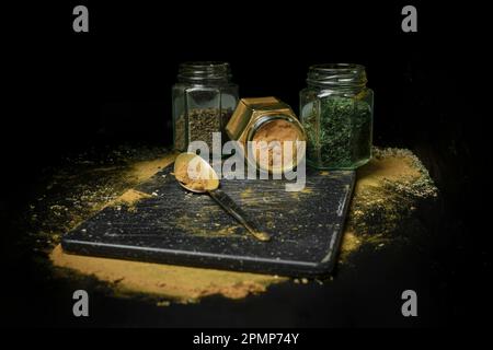 Trois sortes d'herbes ou d'épices dans des bocaux en verre assis dans une rangée sur un fond noir avec une cuillère et des poussières d'herbes et d'épices sur le comptoir Banque D'Images