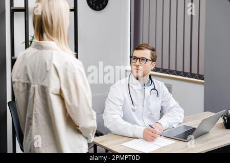 Un jeune médecin attrayant a une consultation dans son bureau. Le médecin donne des recommandations à son patient lorsqu'il est assis sur son lieu de travail. Banque D'Images