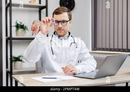 Jeune médecin otolaryngologiste attrayant assis à son lieu de travail dans le bureau et tenant un entonnoir d'oreille dans ses mains. Banque D'Images