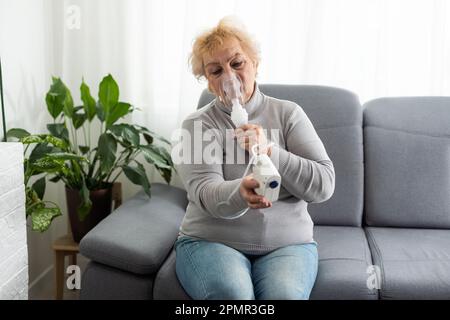 Femme âgée utilisant un nébuliseur fait l'inhalation à la maison et regarde la caméra Banque D'Images