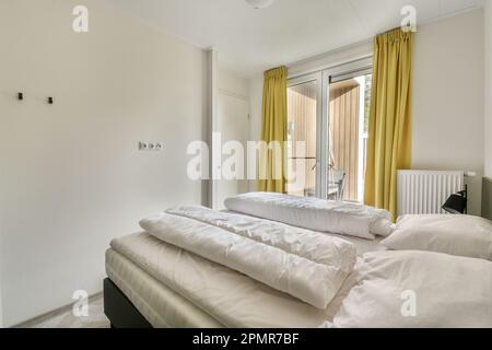 une chambre avec des draps blancs et des rideaux jaunes sur le lit est à côté d'une fenêtre ouverte qui donne sur la rue Banque D'Images