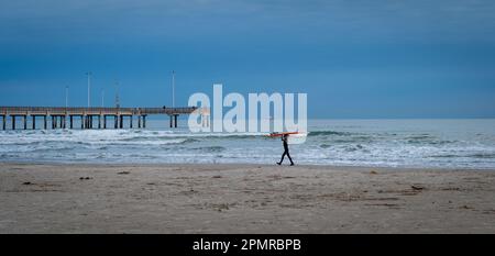 PORT ARANSAS, TX - 9 FEB 2023 : un jeune surfeur porte une planche de surf sur la plage, près de l'eau du golfe du Mexique et d'un quai, à la lumière du soir. Banque D'Images