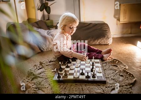 l'enfant joue une seule partie d'échecs à la maison sur le sol.Échiquier avec la fin de la partie d'échecs.La main de la fille se déplaçant une pièce d'échecs est partiellement visible Banque D'Images