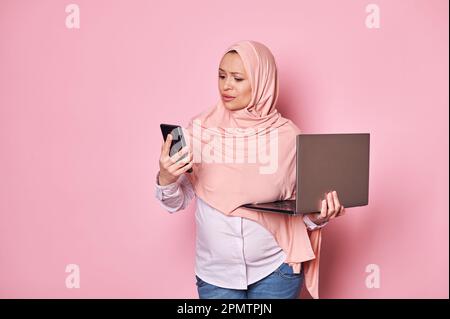 Perplexe femme enceinte arabe musulmane dans le hijab, regardant son smartphone, posant avec un ordinateur portable sur fond rose Banque D'Images