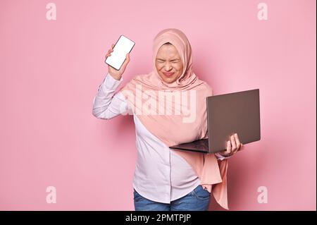 Une femme musulmane enceinte en hijab, avec un smartphone et un ordinateur portable, surtravaillant au bureau, attend avec impatience le congé de maternité Banque D'Images