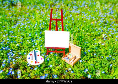 Petite toile vierge sur un petit support en bois, palette miniature et ensemble de peinture nichée dans un champ vert luxuriant. Concept Hello Spring. Banque D'Images