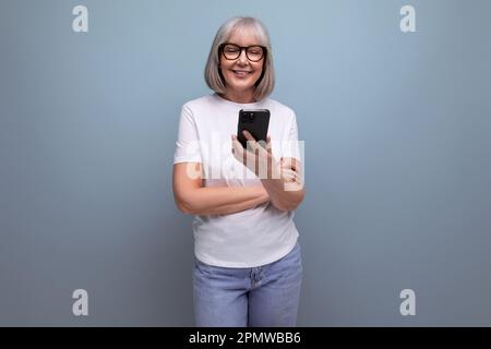 Une femme mûre souriante aux cheveux gris communique sur Internet dans un smartphone sur un arrière-plan studio lumineux Banque D'Images