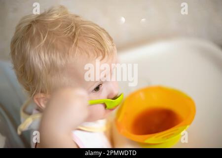 fille de 1 ans, assise sur une chaise pour nourrir et mange une cuillère verte borscht dans une assiette d'enfant orange. enfant qui sait manger seul. Banque D'Images