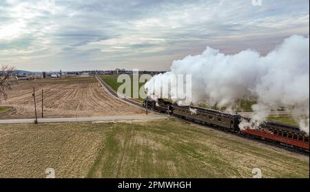 Ronks, Pennsylvanie, 27 décembre 2022 - Une vue aérienne derrière un train de passagers à vapeur qui s'approche, traversant des terres agricoles ouvertes, soufflant beaucoup de fumée blanche, lors d'une journée d'hiver Banque D'Images
