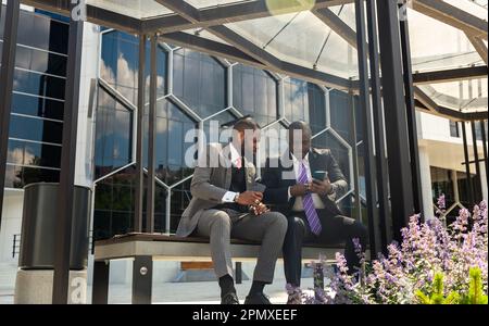 Deux hommes d'affaires à la peau sombre en costume communiquent. Des amis et des partenaires d'affaires sont assis sur un banc dans la ville sur fond de verre Banque D'Images