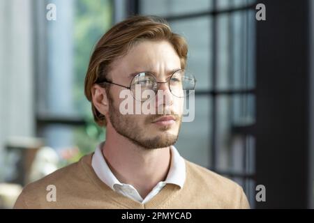 Gros plan. Portrait d'un jeune homme blond en lunettes regardant sérieusement sur le côté, debout dans un bureau, campus, portant une chemise et un chandail brun. Banque D'Images