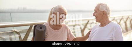 Une femme aîée souriante avec tapis salue un homme avec des bâtons pour la marche nordique sur la passerelle Banque D'Images