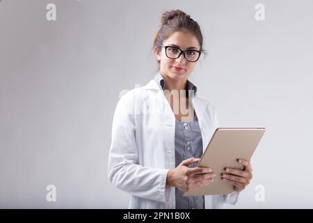 Une femme portant un manteau de laboratoire blanc et des lunettes tient une tablette numérique et regarde directement le spectateur. Est-elle un professionnel de la médecine, un technol Banque D'Images