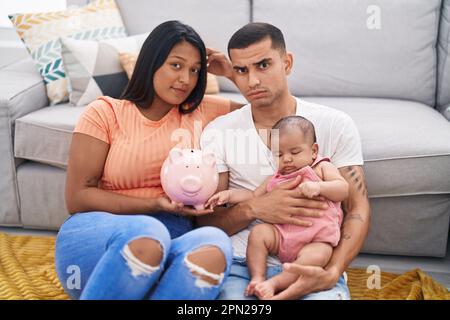 Jeune couple hispanique avec bébé tenant la banque de porc sceptique et nerveux, frowning bouleversé à cause du problème. personne négative. Banque D'Images