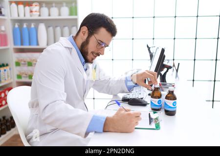 Jeune homme hispanique pharmacien tenant une bouteille de médicament écrit sur le document à la pharmacie Banque D'Images