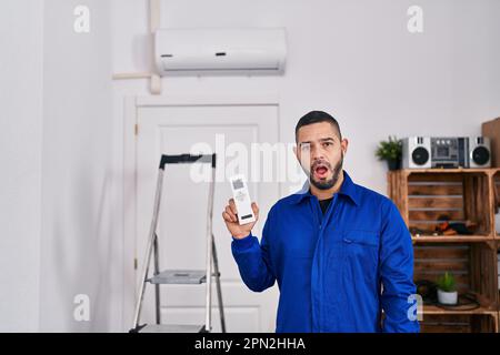 Réparateur hispanique travaillant avec le climatiseur peur et stupéfait avec la bouche ouverte pour la surprise, incrédulité visage Banque D'Images