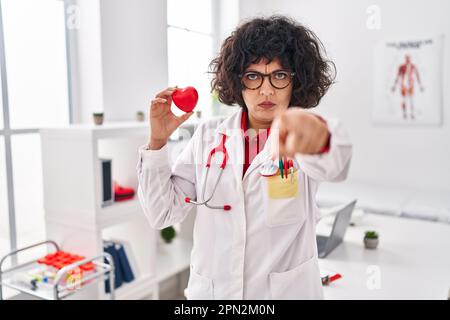 Femme hispanique médecin avec les cheveux bouclés tenant le coeur pointant avec le doigt vers l'appareil photo et à vous, geste confiant regardant sérieux Banque D'Images