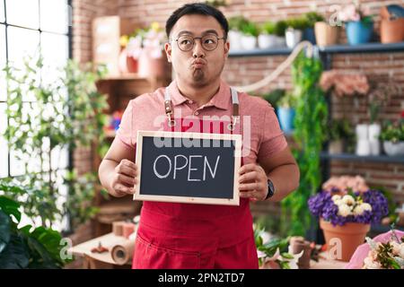 Jeune homme chinois travaillant chez un fleuriste tenant un panneau ouvert faisant le visage de poisson avec la bouche et les yeux accroupidés, fou et comique. Banque D'Images