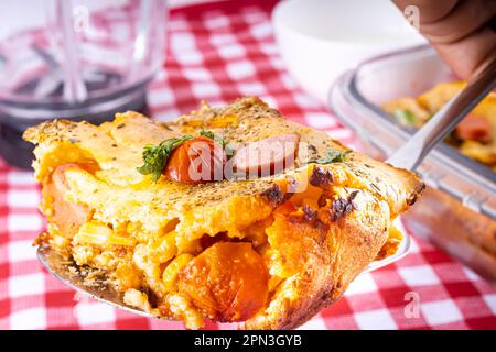 Coupez un morceau de tarte aux saucisses dans un blender, plat typiquement brésilien. Ingrédients : lait, blé, huile, saucisse et maïs. Sur une nappe à carreaux. Banque D'Images