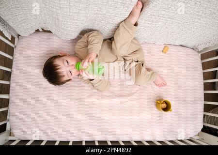 L'enfant mange des biscuits avec du lait provenant d'une bouteille dans un lit de bébé. Nourrir un bébé au lit comme dans un parc. Enfant âgé d'environ deux ans (un an neuf mois) Banque D'Images