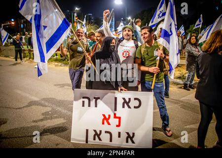 Netanya, Israël. 15th avril 2023. Les manifestants israéliens détenteurs de drapeaux se tiennent derrière un écriteau qui se lit en hébreu : « l'Iran n'est pas là » lors d'une manifestation anti-réforme à Netanya au nord de tel-Aviv. Des centaines de milliers de personnes se sont ralliées pour la semaine droite de 15th samedi contre les plans de réforme judiciaire de la coalition de Netanyahou, un jour après que l'agence de notation Moody's ait abaissé les perspectives économiques d'Israël de positif à stable dans le cadre des propositions très controversées de la coalition visant à brider le pouvoir judiciaire. Crédit : SOPA Images Limited/Alamy Live News Banque D'Images