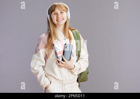 Portrait d'un élève tendance heureux en combinaison beige avec sac à dos, cahier de travail avec drapeau anglais et casque utilisant un smartphone isolé sur fond gris. Banque D'Images