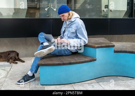 Insouciant gars indépendant travaux types sur ordinateur portable se trouve sur le banc dans la rue dans le parc de la ville. Banque D'Images