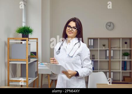 Jeune femme médecin qui pose avec le presse-papiers dans ses mains dans le bureau du centre médical moderne. Banque D'Images