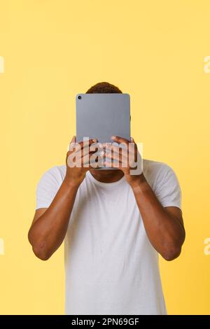 Anonymat incognito en ligne concept. Une personne afro-américaine anonyme tenant un Tablet PC entre les mains, avec une face cachée isolée sur un fond jaune vif avec un espace de copie pour permettre aux concepteurs d'ajouter leur propre texte ou leurs propres graphiques. Photo de haute qualité Banque D'Images